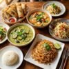 Thai food dishes on wooden table including curry, pad thai, and spring rolls